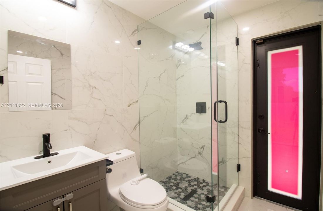 Bathroom featuring large-format veined wall tile, glass-enclosed shower with hexagonal mosaic flooring, dark-finish cabinetry with a white vessel sink, matte black faucet hardware, and a door with a vertical glass accent