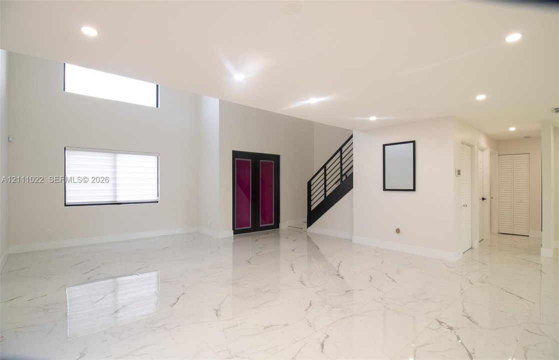 Open-concept interior featuring high ceilings, light-reflective stone-finish flooring, recessed lighting, double-door entryway, and a staircase with a black metal railing