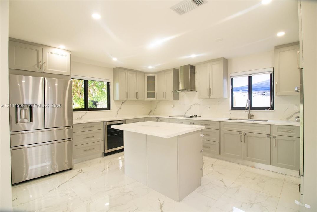 Kitchen featuring light grey shaker-style cabinetry, a center island with a stone-finish countertop, polished tile flooring with veining, stainless steel appliances, and a stainless steel chimney-style range hood