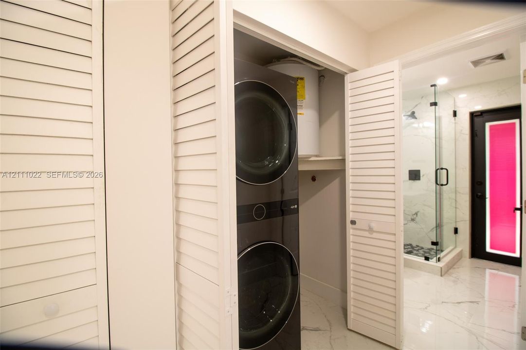 Laundry closet featuring white louvered bifold doors, a stacked washer and dryer unit, and stone-finish flooring extending into the bathroom