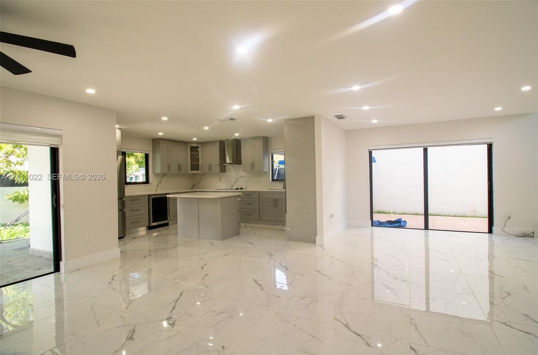 Open-concept space featuring polished stone-finish tile flooring, recessed ceiling lighting, a central kitchen island, grey shaker-style cabinetry, and floor-to-ceiling sliding glass doors