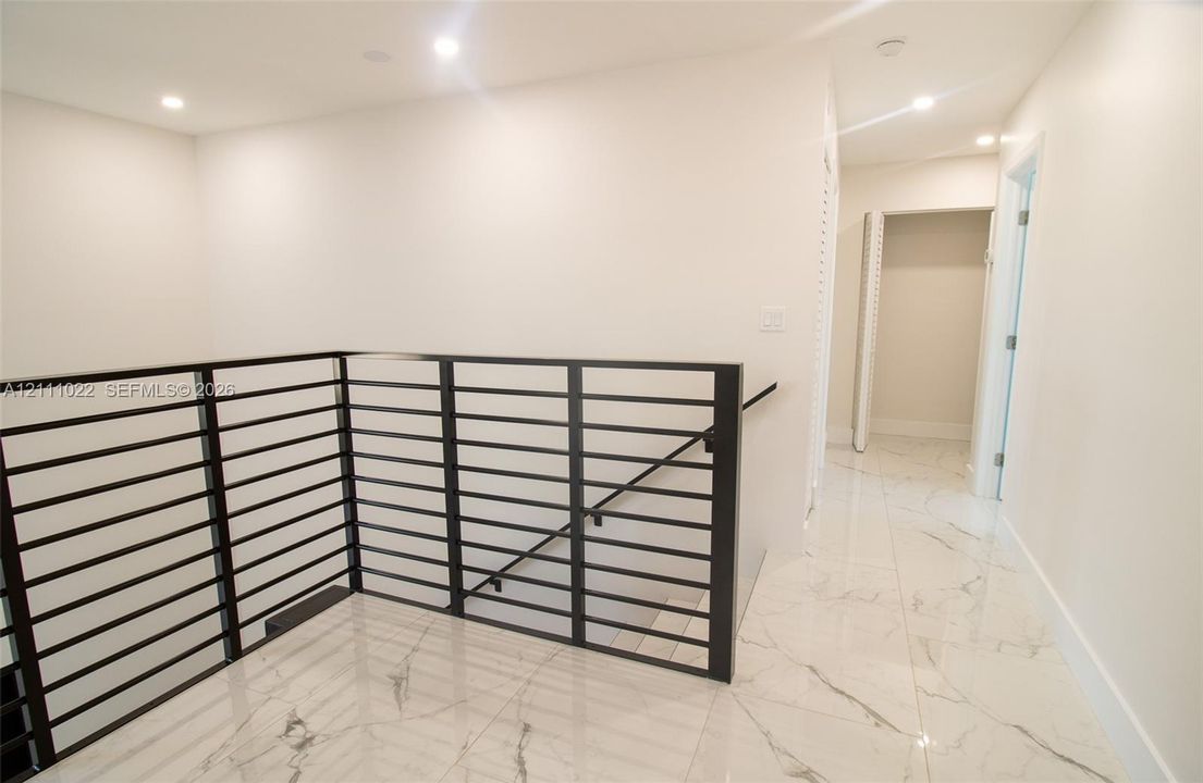 Interior hallway featuring large-format tile flooring with veining detail, recessed lighting, and a metal horizontal-bar railing