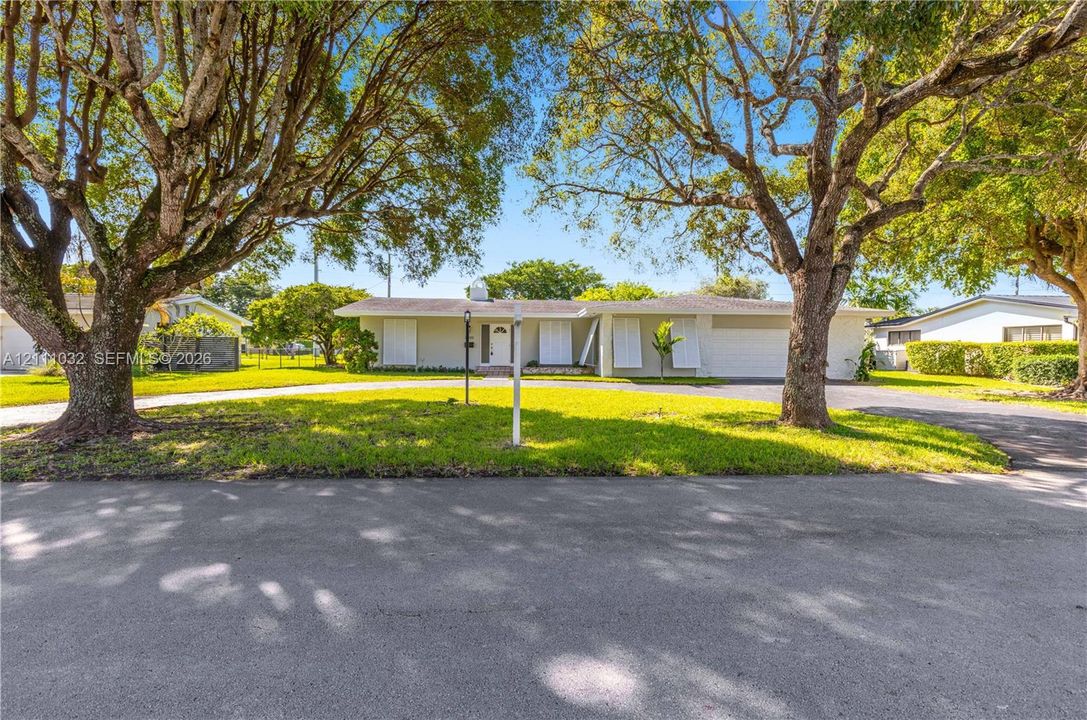 Single-story residence featuring a light-colored stucco exterior, attached two-car garage, white louvered window shutters, and expansive front lawn with mature canopy trees
