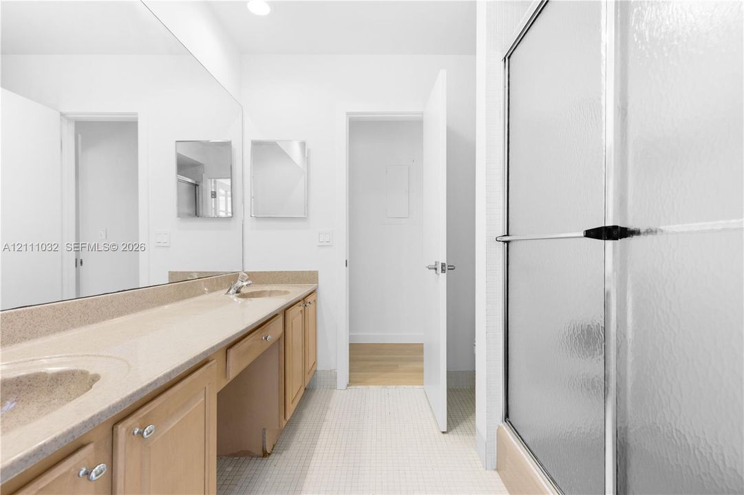 Bathroom featuring a dual-sink vanity with light-tone wood cabinetry, a long wall-to-wall mirror, and a shower enclosure with a frosted glass door