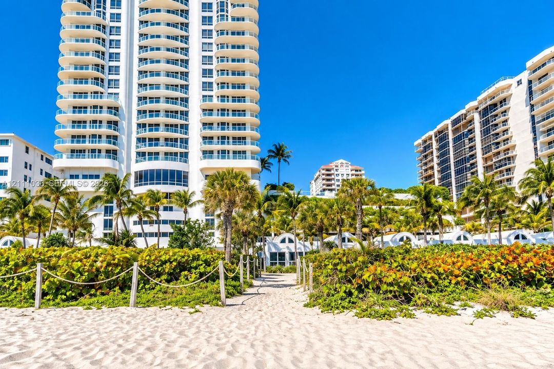 High-rise building featuring curved balconies, white exterior finish, direct sand access, mature palm trees, and rope-lined pedestrian path