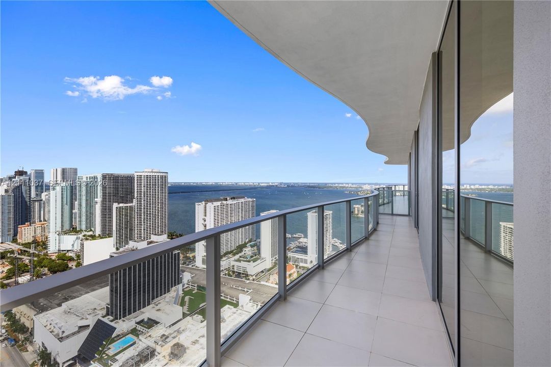 Expansive wrap-around balcony featuring large-format tile flooring, a curved architectural overhang, and floor-to-ceiling glass railings overlooking the water and urban skyline