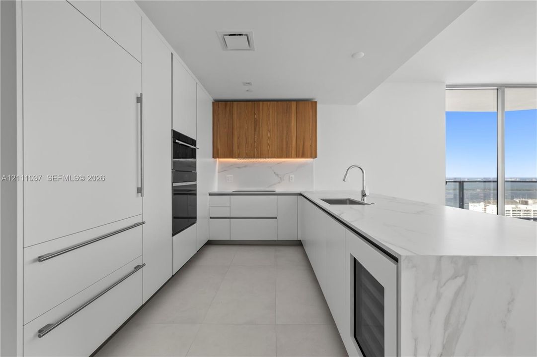 Contemporary kitchen featuring flat-panel white cabinetry, stone countertops with waterfall edge, wood-finish upper cabinets, integrated stainless steel appliances, and large-format tile flooring