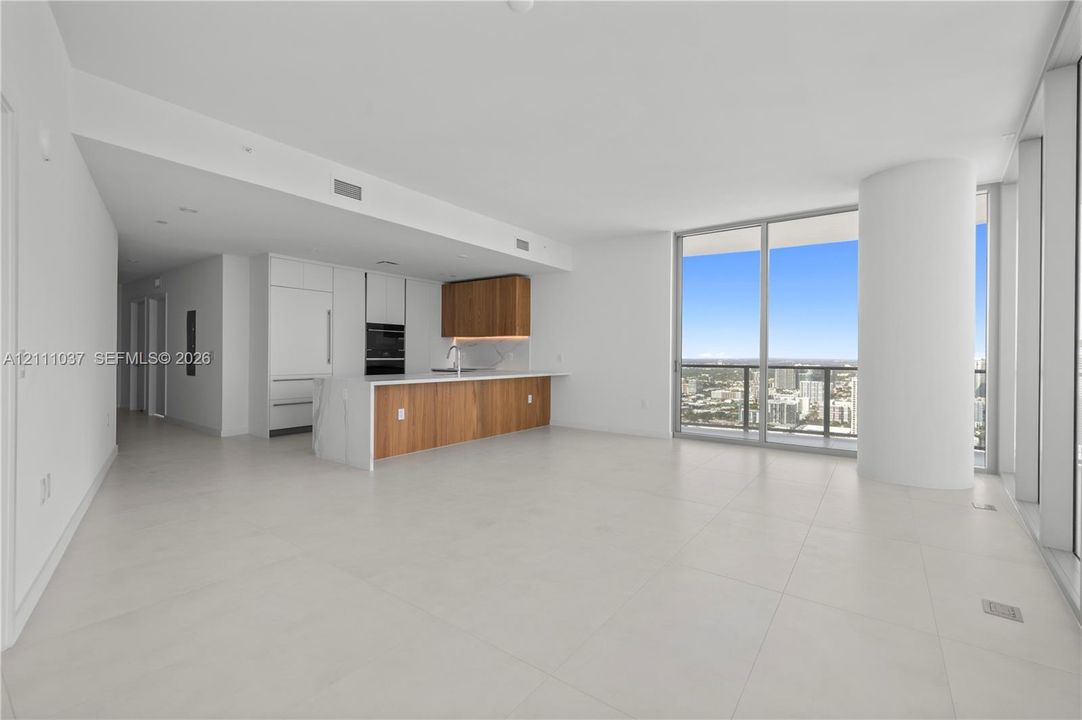 Open-concept interior featuring light-toned tile flooring, floor-to-ceiling windows, and a modern kitchen with wood-finish cabinetry, stone-surfaced waterfall island, and built-in appliances