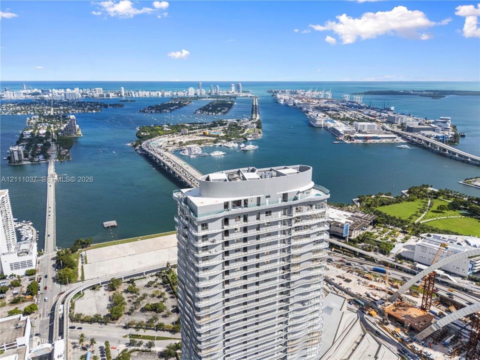 High-rise building featuring glass-railed balconies and a multi-level rooftop terrace, situated in a coastal urban setting with surrounding bay access and multi-lane bridge infrastructure