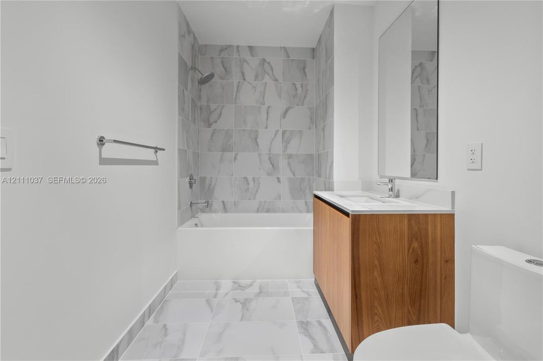 Bathroom featuring a wood-finish vanity with a white countertop, large-format tile flooring, a tub-and-shower combination with floor-to-ceiling surround tile, and a wall-mounted mirror