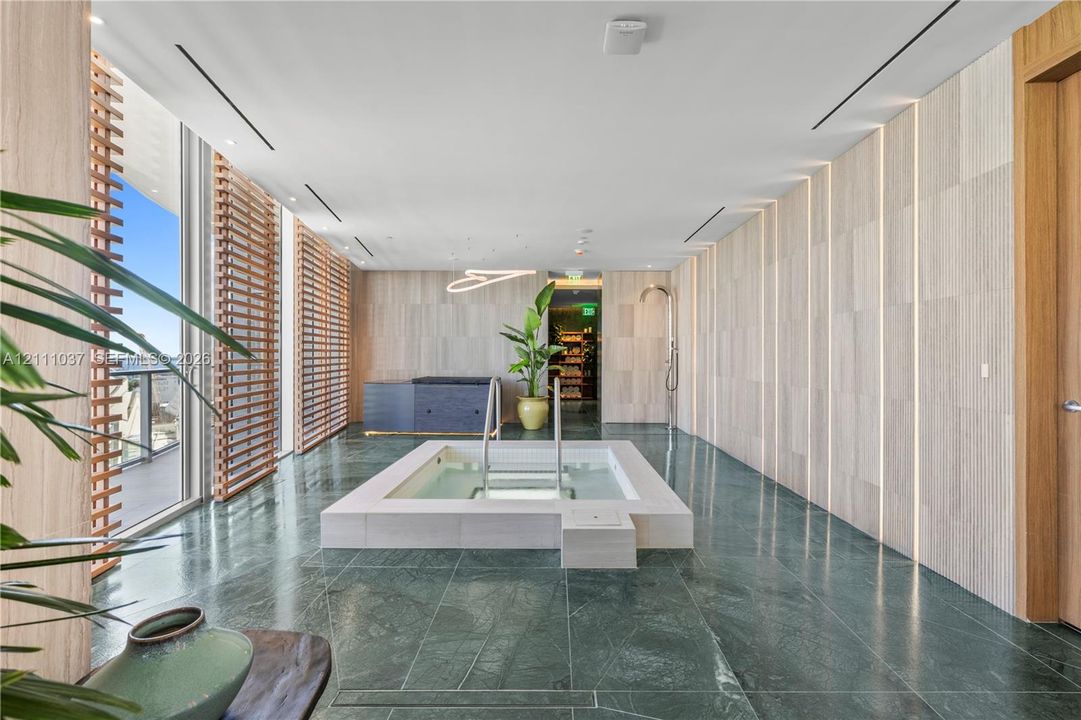 Spa-inspired room featuring a central sunken jetted tub, stone-tile flooring, floor-to-ceiling textured wall paneling with recessed linear lighting, and wooden louvered window screens