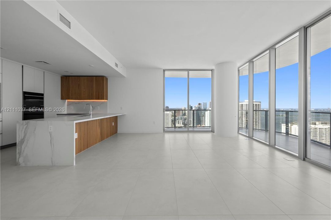 Open-concept space featuring floor-to-ceiling glass walls, large-format tile flooring, a kitchen island with stone-finish waterfall countertop, and custom wood-finish cabinetry