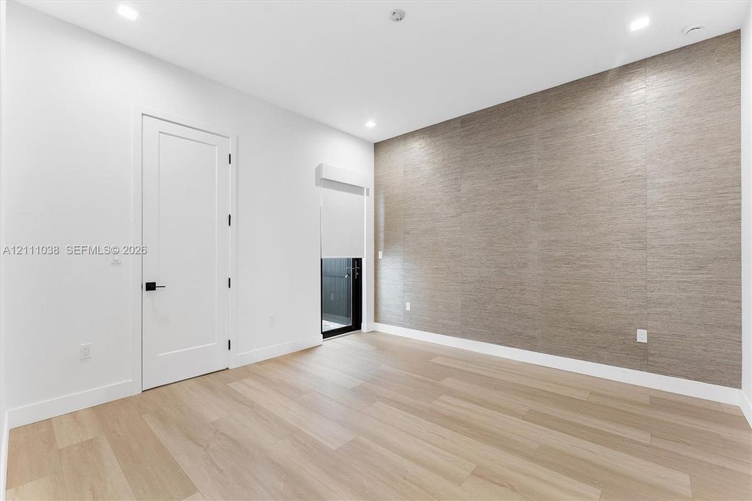 Interior space featuring a textured accent wall, light-toned wood-finish flooring, recessed ceiling lighting, a solid-core interior door with matte black hardware, and a window fitted with a roller shade