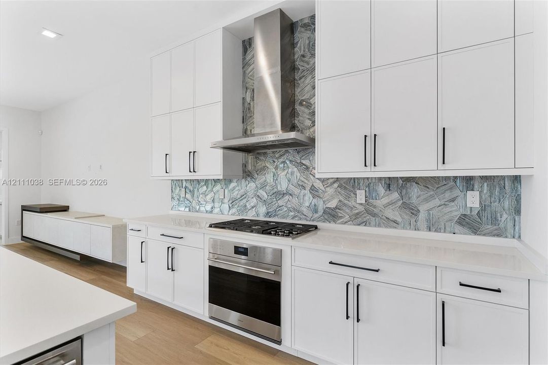 Kitchen featuring white cabinetry with matte black hardware, stone-finish countertops, stainless steel gas range with wall-mounted vent hood, and multi-tone geometric tile backsplash