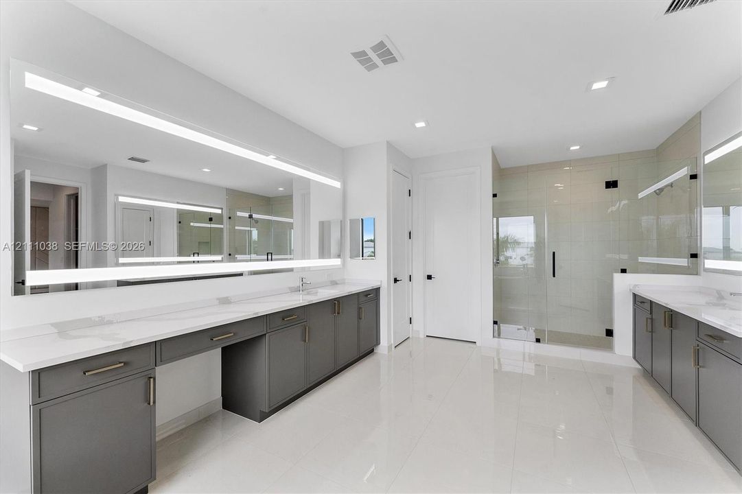 Dual vanities featuring charcoal cabinetry and white stone countertops, expansive wall-to-wall LED-backlit mirrors, polished tile flooring, and a glass-enclosed shower with tile surround