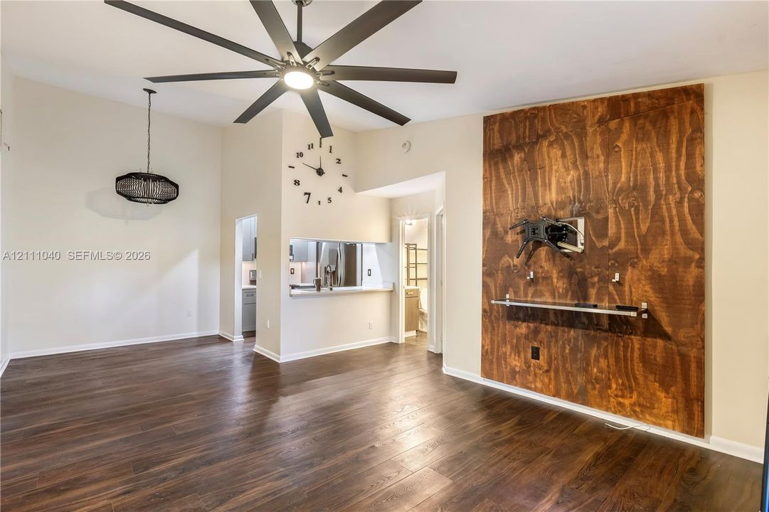 Vaulted ceiling featuring a multi-blade matte black ceiling fan, wood-finish flooring throughout, and a custom wood-paneled accent wall with an attached glass shelving unit