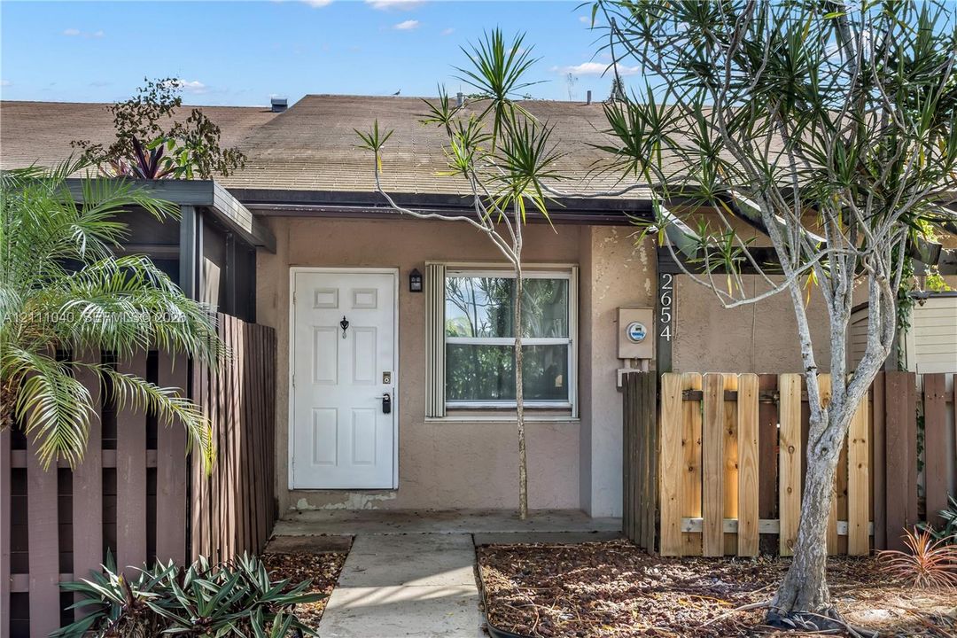 Exterior entrance featuring a concrete walkway, six-panel entry door, single-hung window with framing, and a wood-slat privacy fence