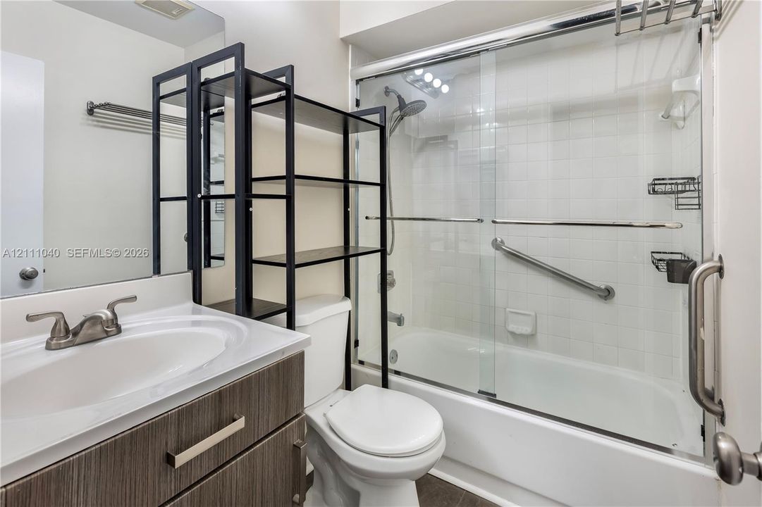 Bathroom vanity with a neutral-toned countertop and wood-finish cabinetry, complemented by a shower-tub combination enclosed by sliding glass doors, polished metal hardware, and wall-mounted grab bars