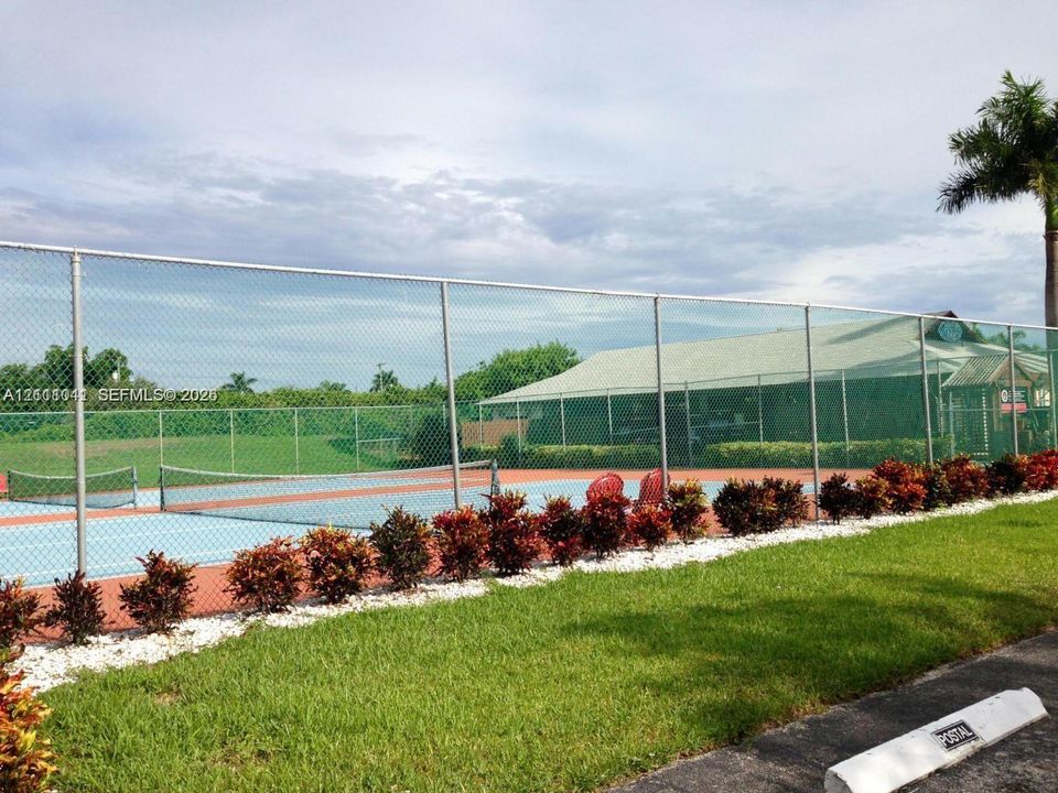 Hard-surface tennis court enclosed by chain-link fencing, featuring professional netting and manicured landscaping with white gravel border