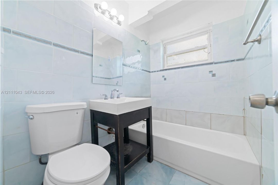 Bathroom featuring light blue wall tiles with decorative border accents, dark-finish pedestal vanity, integrated white sink, and combination bathtub with shower fixture