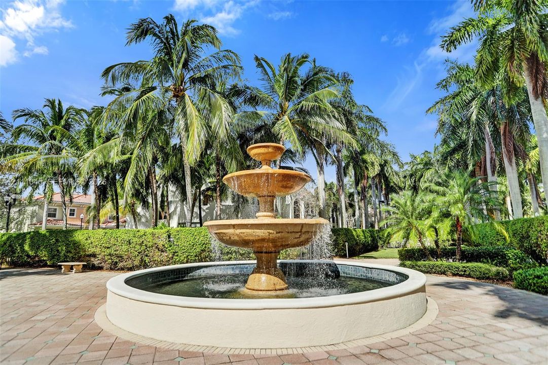 Three-tiered stone fountain centerpiece, circular paved plaza, and lush perimeter hedges framed by mature palm trees