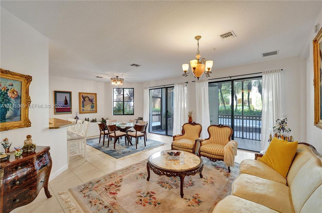 Open-concept living space featuring large-format tile flooring, dual multi-light chandeliers, built-in breakfast bar with stone-finish countertop, and floor-to-ceiling sliding glass doors leading to a screened exterior area