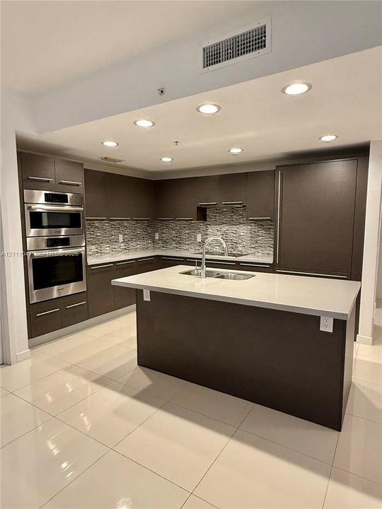 Kitchen featuring flat-panel cabinetry, a central island with a double-basin sink, stone countertops, a multi-tonal tile backsplash, and built-in stainless steel appliances