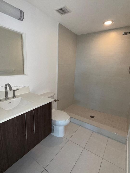 Bathroom featuring a dark wood-finish vanity with a white countertop and rectangular vessel sink, large-format tiled walk-in shower with a chrome rainfall showerhead, and light-toned ceramic tile flooring