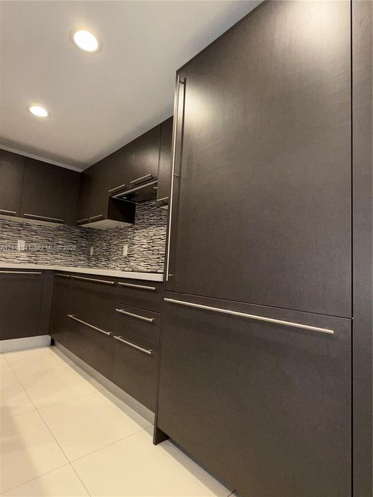 Flat-panel cabinetry in a dark finish with brushed metallic hardware, integrated refrigerator, multi-toned stone mosaic tile backsplash, and light-toned tile flooring