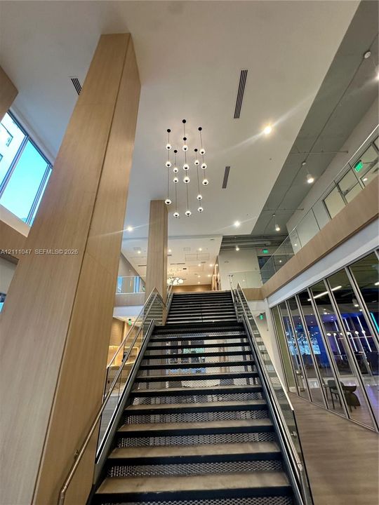 Double-height ceiling featuring wood-paneled pillars, suspended multi-globe chandelier, metal staircase with glass railing, and floor-to-ceiling glass wall partitions