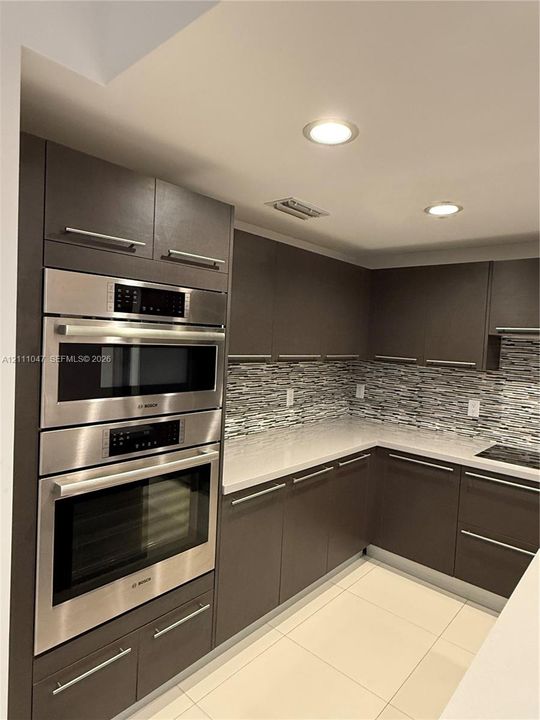 Kitchen featuring charcoal flat-panel cabinetry, light-toned stone countertops, built-in stainless steel Bosch oven and microwave, mosaic tile backsplash, and tile flooring