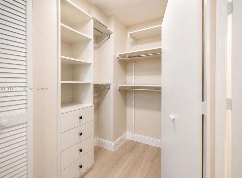 Walk-in closet featuring custom built-in shelving, integrated four-drawer storage, polished metal hanging rods, wood-finish flooring, and a white louvered door
