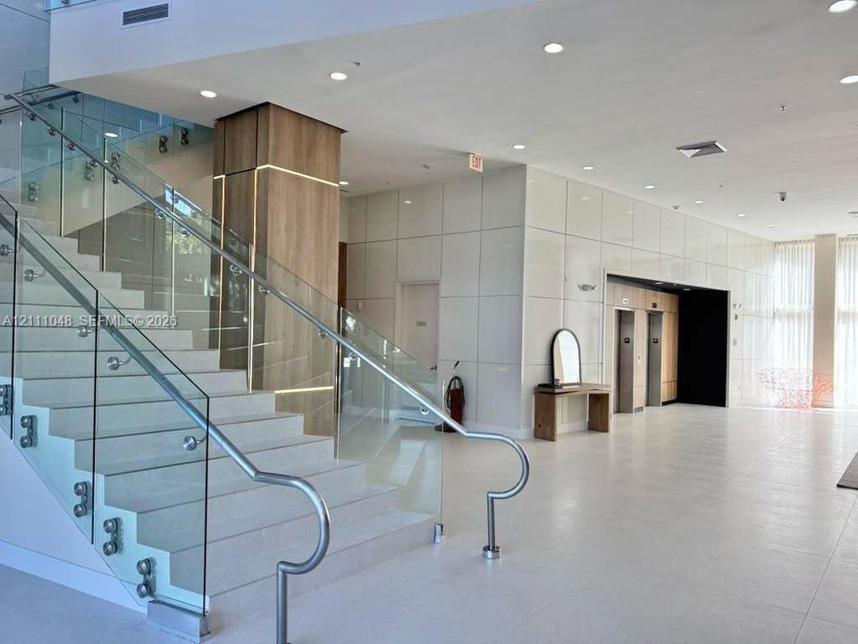 Lobby area featuring a wide staircase with glass railings and metal handrails, a central pillar with integrated accent lighting and wood-finish paneling, large-format tiled flooring, and high-gloss wall panels