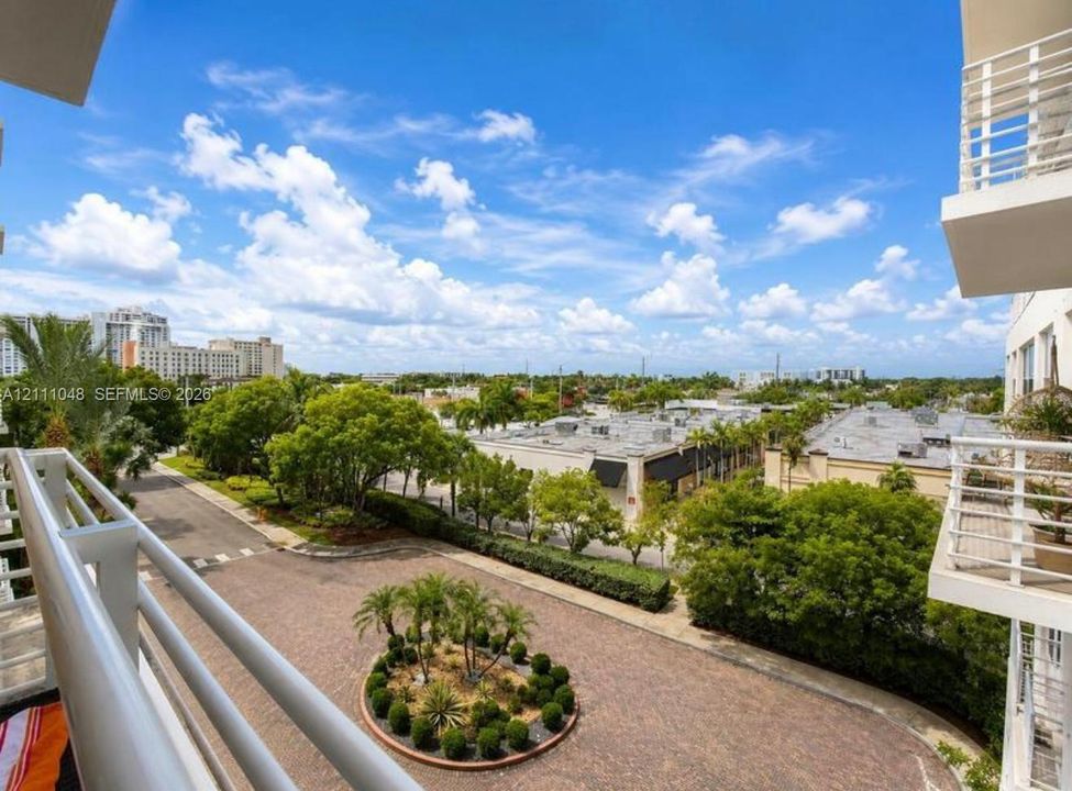 Elevated balcony with white metal railings overlooking a paved circular drive featuring manicured landscaping and mature trees