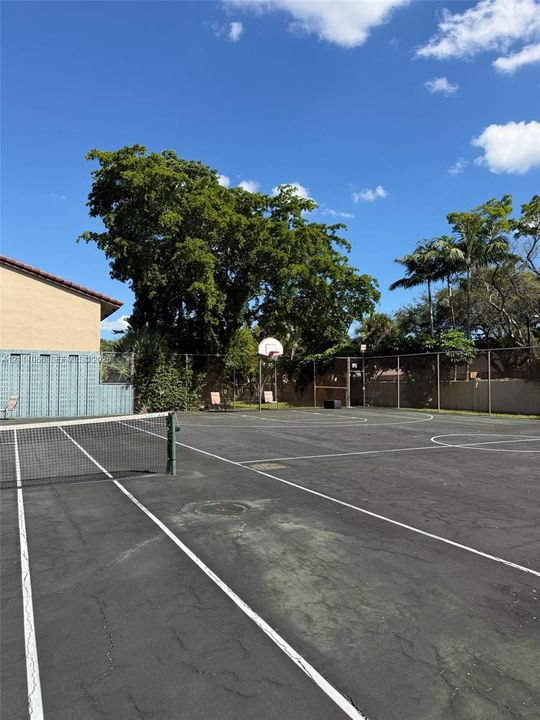 Multi-purpose recreation court featuring a basketball hoop, tennis net, perimeter fencing, and mature landscaping