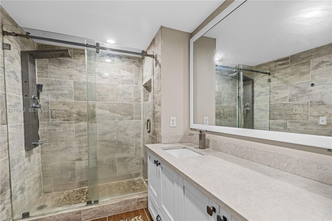 Bathroom featuring a walk-in shower with sliding glass doors, stone-finish wall tile, a built-in shower panel, white cabinetry with a stone-finish countertop, and a large framed vanity mirror