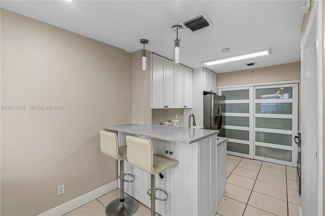 Kitchen featuring white shaker-style cabinetry, stone-surface breakfast bar with integrated sink, stainless steel refrigerator, and frosted-glass panel sliding doors, finished with tile flooring and dual pendant lighting