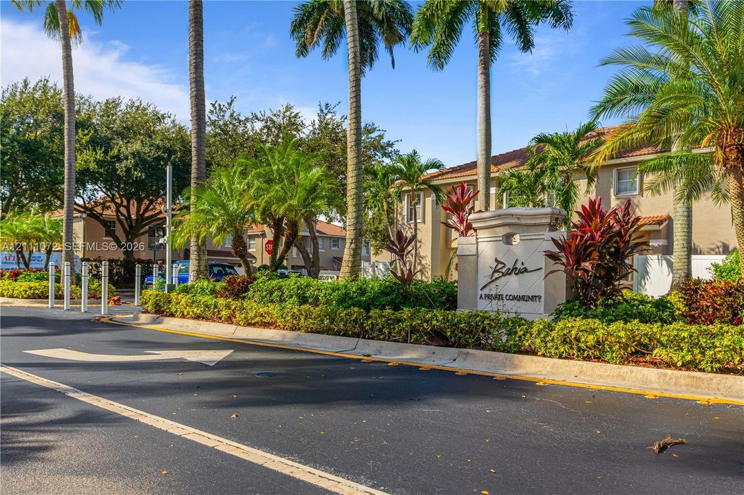 Community entrance featuring mature palm trees, manicured hedges, colorful tropical shrubbery, and decorative stone-finish signage