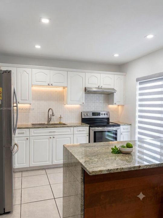 Kitchen featuring white cabinetry, stone-surface countertops, tile backsplash with under-cabinet lighting, stainless steel appliances, and a center island with a wood-finish base
