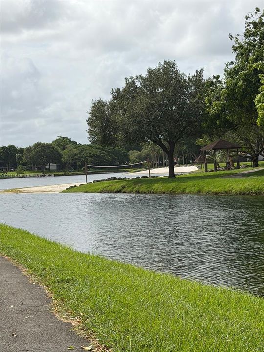 Waterfront grounds featuring a sandy shoreline, paved perimeter path, mature trees, and a wooden gazebo