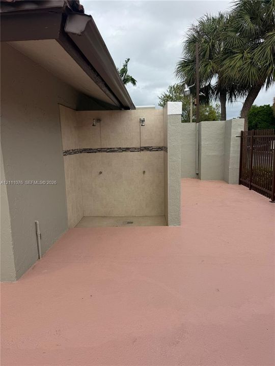 Outdoor shower with dual showerheads, stone-tiled wall finish, and decorative accent border, complemented by an expansive concrete patio area and privacy wall enclosure