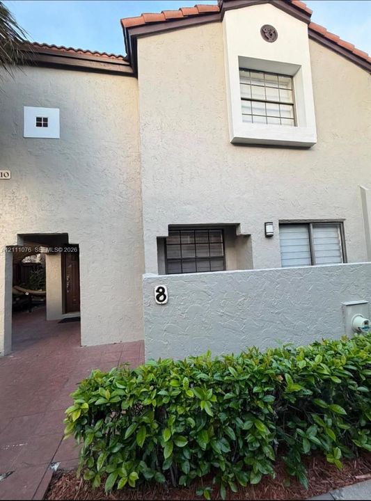 Stucco exterior with a tiled roof, recessed entryway, masonry privacy wall, and manicured shrubbery