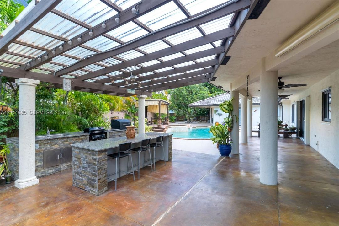 Covered patio featuring an outdoor kitchen with stone veneer base, countertop bar seating, a slatted timber pergola, and a concrete pool deck