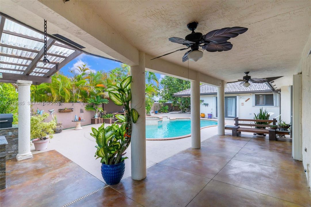 Covered patio with stained concrete flooring, supported by round columns, featuring dual ceiling fans and a built-in stone-veneer counter, overlooking an in-ground swimming pool