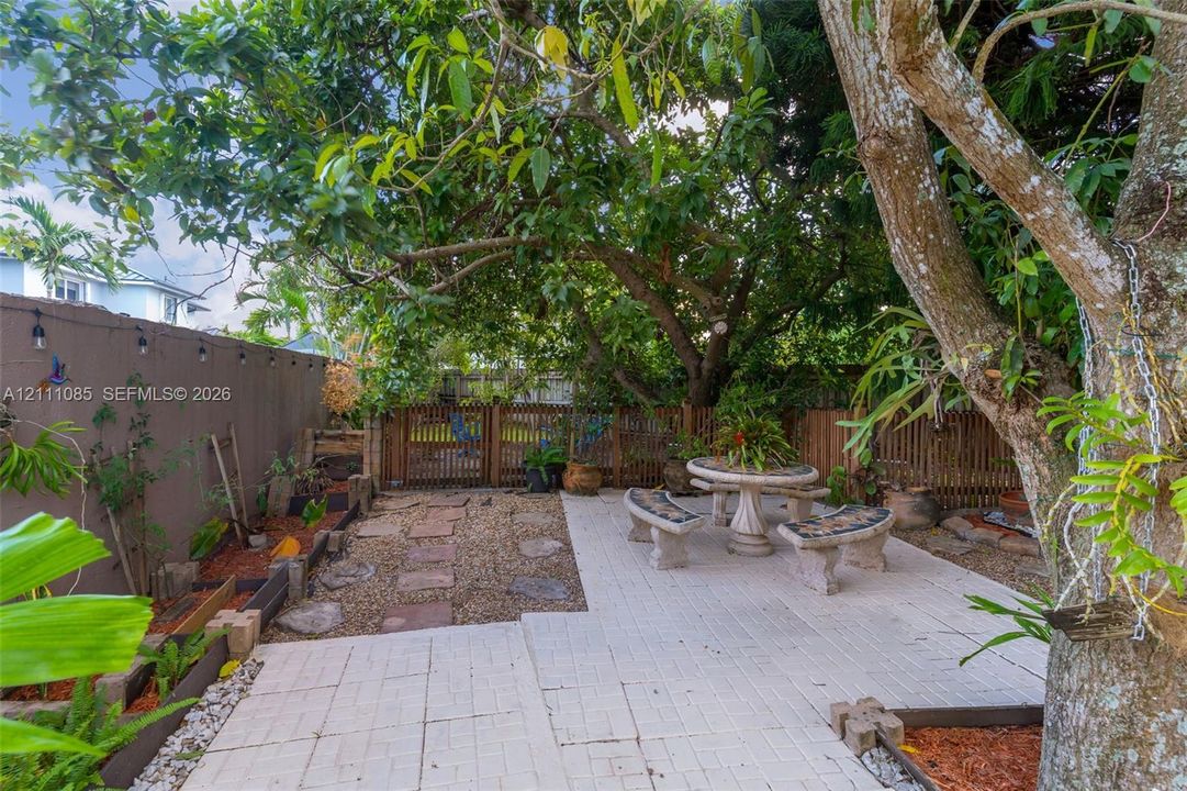 Outdoor space featuring a light-toned paver patio, stone-finish built-in seating, a gravel garden path, mature trees, and a perimeter wall with wall-mounted lighting