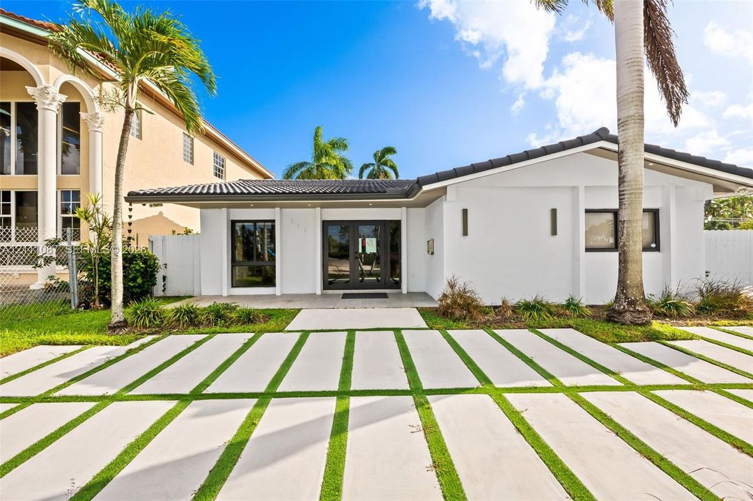 Single-story exterior featuring white stucco siding, a tiled roof, dual-entry glass doors, and an expansive concrete paver driveway with integrated grass turf