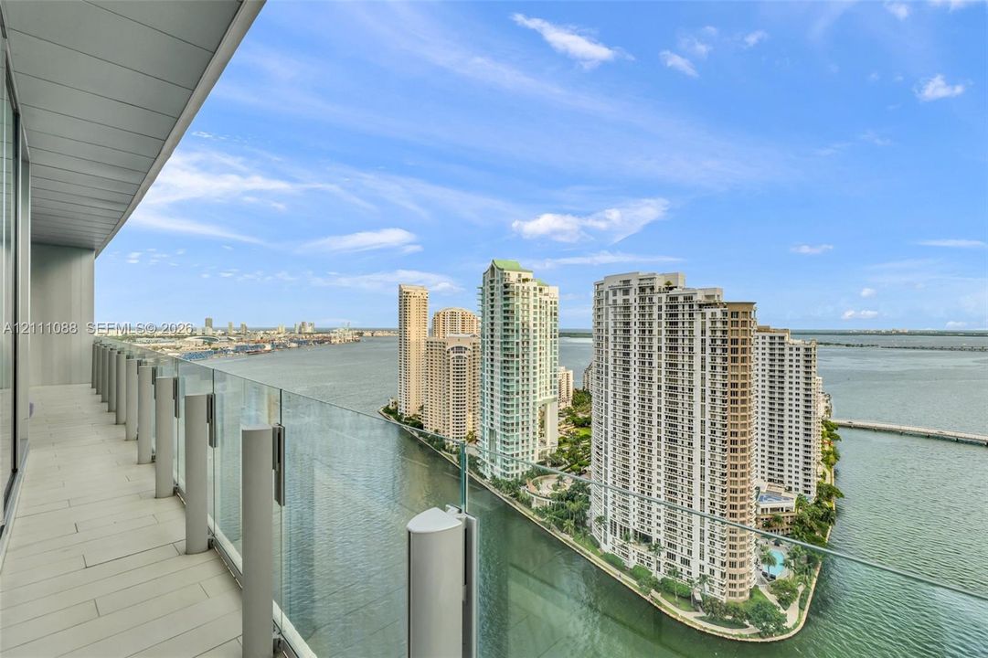 Covered balcony with light-toned flooring, frameless glass balustrades, and expansive water and urban skyline scenery