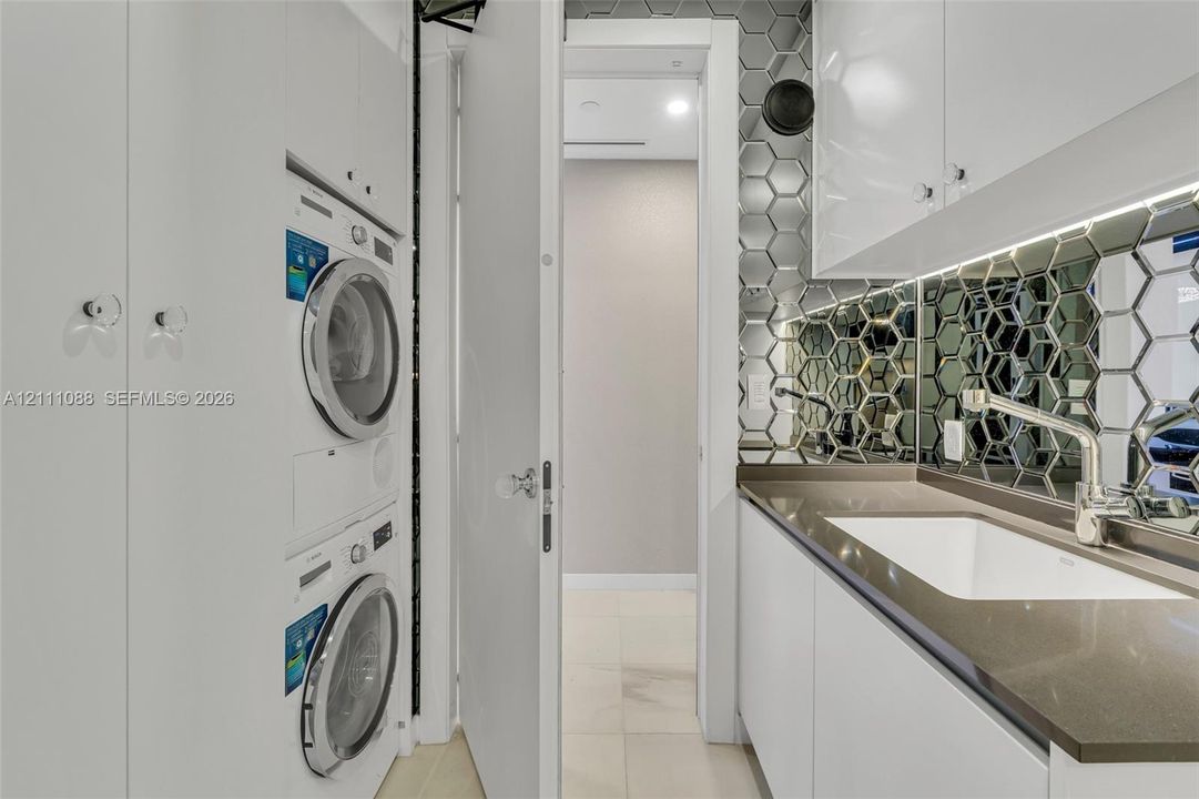 Utility space featuring high-gloss white cabinetry, integrated stacking laundry appliances, stone-finish countertops with an under-mount sink, and a metallic hexagonal tile backsplash
