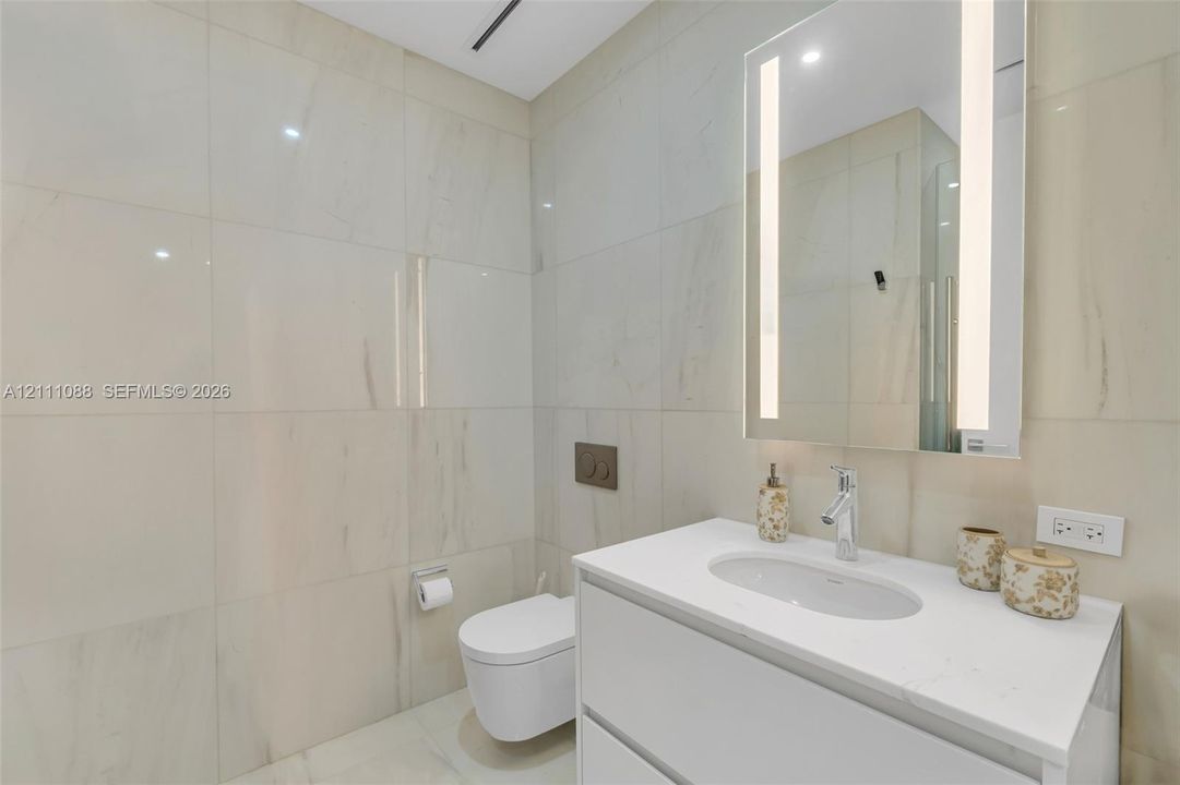 Full-height wall tiling, wall-mounted commode with a dual-flush actuator plate, floating vanity with a white countertop and integrated sink, and an LED-integrated vanity mirror
