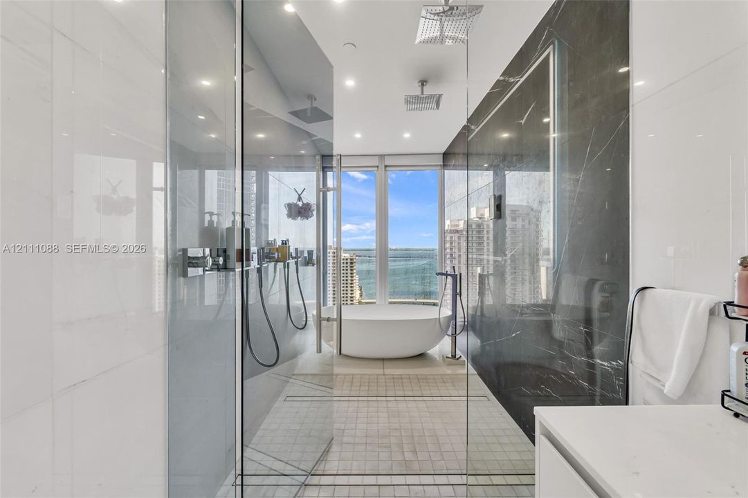 Walk-in glass-enclosed shower featuring dual rain-style showerheads and handheld fixtures, freestanding soaking tub, contrasting large-format wall tiling, and floor-to-ceiling windows